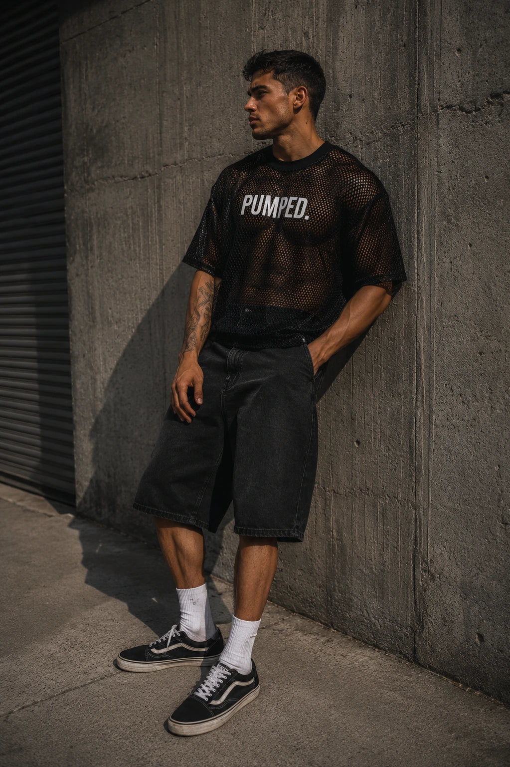 Man wearing a black 'PUMPED.' t-shirt leaning against a concrete wall.